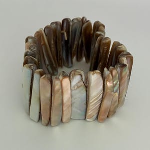 May include: A stretch bracelet composed of numerous vertical, rectangular pieces of iridescent shell. The segments display a gradient of colors ranging from creamy white and soft peach to deep brown, featuring a natural, polished, and pearlescent finish.