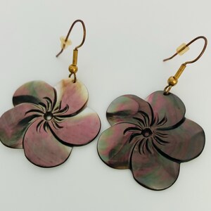 May include: A pair of dangle earrings featuring carved flower-shaped pendants made from iridescent shell material with swirling hues of dark pink, green, and black. The floral charms are attached to gold-toned metal fishhook wires with clear backings.