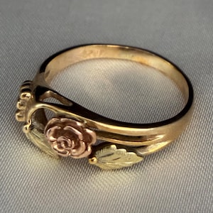 May include: A ring featuring a central rose motif in rose gold, flanked by two textured yellow gold leaves. The band is crafted from polished yellow gold, creating a multi-toned metal design that highlights the floral elements and leaf details.