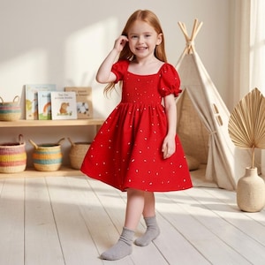 May include: A bright red dress featuring short puff sleeves, a square neckline, and a smocked bodice. The skirt is flared and decorated with a scattered pattern of small white dashes. The dress is worn with simple grey ankle socks.