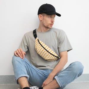 Könnte beinhalten: Eine Bauchtasche mit cremefarbener Basis und einem sich wiederholenden Muster aus senfgelben Punkten, ausgestattet mit einem schwarzen Reißverschluss und einem verstellbaren schwarzen Gurt mit Kunststoffschnalle, getragen über einem grauen T-Shirt.