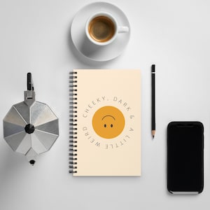 May include: A spiral-bound notebook with a cream-colored cover featuring a central yellow circle with an upside-down smiley face and the text "CHEEKY, DARK & A LITTLE WEIRD" arranged in a circular pattern around the graphic.