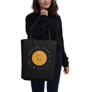 May include: A black tote bag featuring a central yellow circle with an upside-down smiley face. The text "CHEEKY, DARK & A LITTLE WEIRD" is printed in a circular white font around the yellow graphic, with two black shoulder straps.