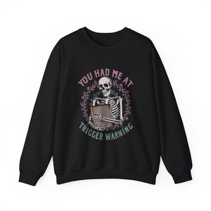 Puede incluir: Una sudadera negra de cuello redondo con un gráfico de un esqueleto sosteniendo un libro, rodeado por una corona floral. El texto "YOU HAD ME AT TRIGGER WARNING" aparece en letras rosas y verdes en el centro del pecho de la prenda.