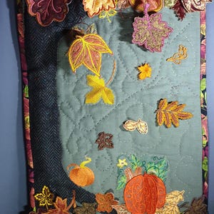 May include: A rectangular quilted wall hanging featuring a sage green center panel with embroidered autumn leaves and pumpkins. The border consists of dark herringbone fabric and a colorful leaf-patterned trim, with three-dimensional fabric leaves.
