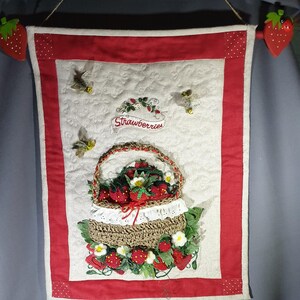 May include: A quilted wall hanging featuring a central cream-colored panel with a red border and white polka-dot corners. It displays a woven basket filled with red strawberries, white flowers, and green leaves, with the text "Strawberries" above.