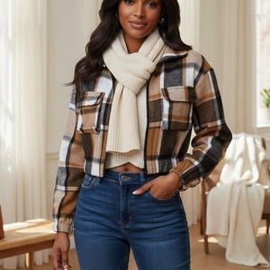 Könnte beinhalten: Eine kurz geschnittene Karo-Jacke mit einem Muster aus braunen, schwarzen und weißen Quadraten, kombiniert mit einem cremefarbenen Rippstrickschal und einer dunkelblauen Jeans. Die Jacke hat einen Frontreißverschluss und zwei Brusttaschen mit Klappen.