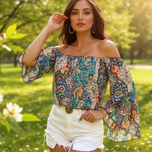 May include: An off-the-shoulder blouse featuring a vibrant, multi-coloured paisley pattern with bell sleeves and black trim, paired with white denim shorts that have a frayed hem and a brown circular belt buckle at the waist.