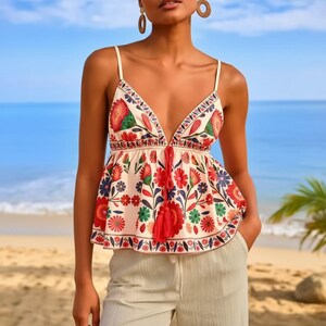 Puede incluir: Un top tipo camisola de color crema con un estampado floral vibrante en tonos rojos, verdes y azules. La prenda tiene tirantes finos, un escote en V profundo y un detalle de cordón con borlas rojas en la cintura, combinado con pantalones beige.