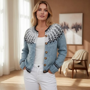 May include: A blue knit cardigan featuring a round neckline and a white yoke with a black geometric pattern. The garment has long sleeves with ribbed cuffs and a front button closure with brown buttons, worn over a plain white top and white pants.