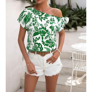 Puede incluir: Una blusa blanca de manga corta con estampado floral verde, escote descubierto y detalle de lazo lateral. Se combina con pantalones cortos de mezclilla blancos con dobladillo deshilachado y un cinturón trenzado marrón con hebilla circular.
