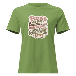 May include: A sage green short-sleeved t-shirt featuring a graphic print with the text "Please do not contact me unless you are an iced matcha latte." The design includes a small illustration of a drink cup, stars, and hearts in pink and brown.