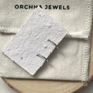 Può includere: Due orecchini a cerchio d'argento su uno sfondo di tessuto bianco con il testo "ORCHHA JEWELS" in nero.