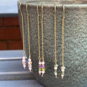 May include: Five pairs of gold-coloured dangle earrings with various gemstone beads. The earrings hang from gold-coloured hooks and feature a delicate chain design. The gemstones include pink, green, and purple beads.
