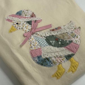 May include: A cream-colored crewneck sweatshirt featuring a large, central appliqué of a duck. The duck is constructed from various patterned fabric patches, including floral and gingham prints, accented with white lace trim and a pink gingham bow.