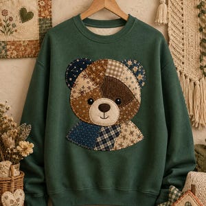 May include: A dark green crewneck sweatshirt featuring a large, central applique of a teddy bear face. The bear is constructed from various fabric patches in brown, blue, and cream, showcasing floral, plaid, and polka dot patterns with a stitched finish.