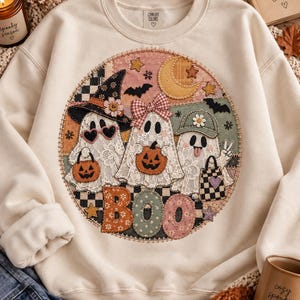 May include: A cream-colored crewneck sweatshirt featuring a large circular patch design. The graphic displays three ghosts with lace details, wearing hats and accessories, holding pumpkins and a bag. The word BOO is printed in large, patterned letters.