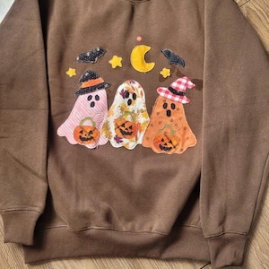 May include: A brown crewneck sweatshirt featuring a central applique design of three ghosts wearing hats and holding jack-o'-lanterns. The scene includes a yellow crescent moon, small yellow stars, and two black bats against the brown fabric.