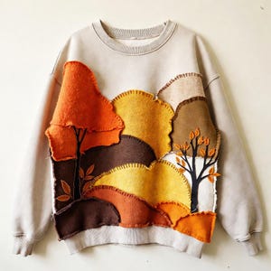 May include: A beige crewneck sweatshirt featuring a large, layered fabric appliqué on the front. The design depicts a landscape of rolling hills and trees in shades of orange, yellow, and brown, with visible blanket stitching along the edges.