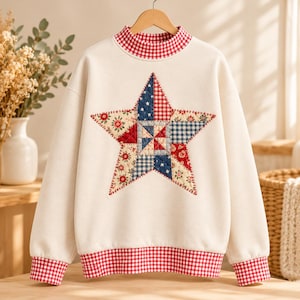 May include: A cream-colored long-sleeve sweatshirt featuring a large, quilted star applique on the chest. The star is composed of various fabric patches in red, blue, and white floral and gingham patterns. The collar and cuffs have red gingham.