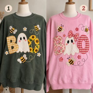 May include: Two crewneck sweatshirts displayed on mannequins. The dark green sweatshirt features a white ghost, yellow gingham 'B', sunflower 'O', and bees. The pink sweatshirt features a white ghost with a floral bow, floral 'B', red gingham 'O', and bees.