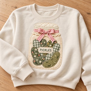 May include: A cream-colored crewneck sweatshirt featuring a large, quilted applique of a glass jar filled with fabric pickles. The jar is decorated with a pink and white gingham ribbon bow and lace trim, with the word PICKLES printed on a label.