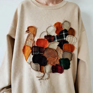 May include: A beige crewneck sweatshirt featuring a large, central appliqué of a turkey. The turkey is constructed from various fabric patches in shades of orange, brown, cream, green, and black, including plaid and textured patterns with visible stitching.