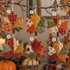 May include: Six fabric maple leaf ornaments numbered one through six, each featuring a patchwork design of various autumn-colored cotton prints including plaid, floral, and solid patterns with visible stitching and twine loops for hanging.