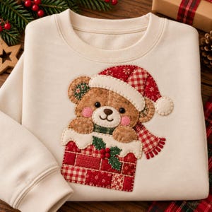 May include: A cream-colored crewneck sweatshirt featuring a large, central fabric appliqué of a brown teddy bear wearing a red and white plaid Santa hat and scarf, peeking out from a red and white patchwork chimney decorated with green holly.