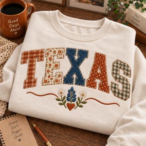 May include: A cream-colored crewneck sweatshirt featuring the word TEXAS in large, appliqued letters. Each letter has a unique pattern, including plaid, floral, denim, and polka dot fabrics. Below the text is an embroidered floral design.