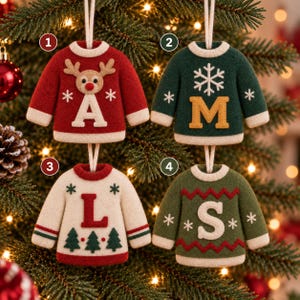 May include: Four felt sweater ornaments hanging by white strings. Ornament 1 is red with a reindeer and the letter A. Ornament 2 is dark green with a white snowflake and the letter M. Ornament 3 is cream with a red L and trees. Ornament 4 is olive green with a white S.