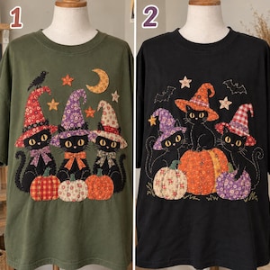 May include: Two t-shirts displayed on mannequins. The olive green shirt features three black cats wearing patterned witch hats and bows, sitting by pumpkins with a moon and stars. The black shirt shows three black cats with pumpkins and bats.