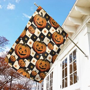 May include: A rectangular garden flag featuring a repeating pattern of orange jack-o'-lanterns with carved faces set against a black and cream checkered background, accented with small gold stars, mounted on a metal pole attached to a white wall.