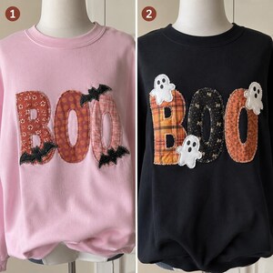 May include: Two crewneck sweatshirts displayed on mannequins. The left is light pink with fabric-appliqued letters spelling BOO in orange patterns, accented by black bats. The right is black with orange and black patterned letters and white ghosts.