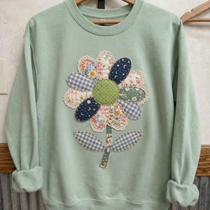 May include: A light sage green crewneck sweatshirt featuring a large, central flower appliqué. The flower has a green polka-dot center surrounded by petals made of various patterned fabrics, including blue gingham, floral prints, and navy with moon motifs.