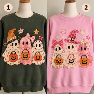 May include: Two crewneck sweatshirts, one dark olive green and one light pink, each featuring a central appliqué design of three ghosts wearing patterned fabric hats and bows, holding orange jack-o'-lantern buckets, accented by small fabric stars.