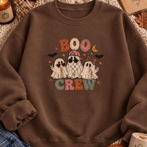 May include: A brown crewneck sweatshirt featuring a graphic of three ghosts with the text "BOO CREW". The ghosts wear accessories including heart-shaped sunglasses, a baseball cap, and a bow. The design includes small stars, bats, and a crescent moon.