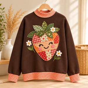 May include: A brown long-sleeve sweatshirt featuring a patchwork strawberry appliqué with a smiling face. The garment has a mock neck, cuffs, and a hemline in a red and white gingham pattern. Two small white flowers are stitched on the sides.