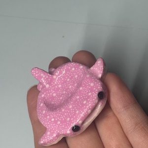 May include: A small, sculpted whale shark figurine featuring a glossy pink surface covered in a pattern of tiny white dots. The creature has two prominent black bead-like eyes and a wide, slightly open mouth, with fins extending from its sides.