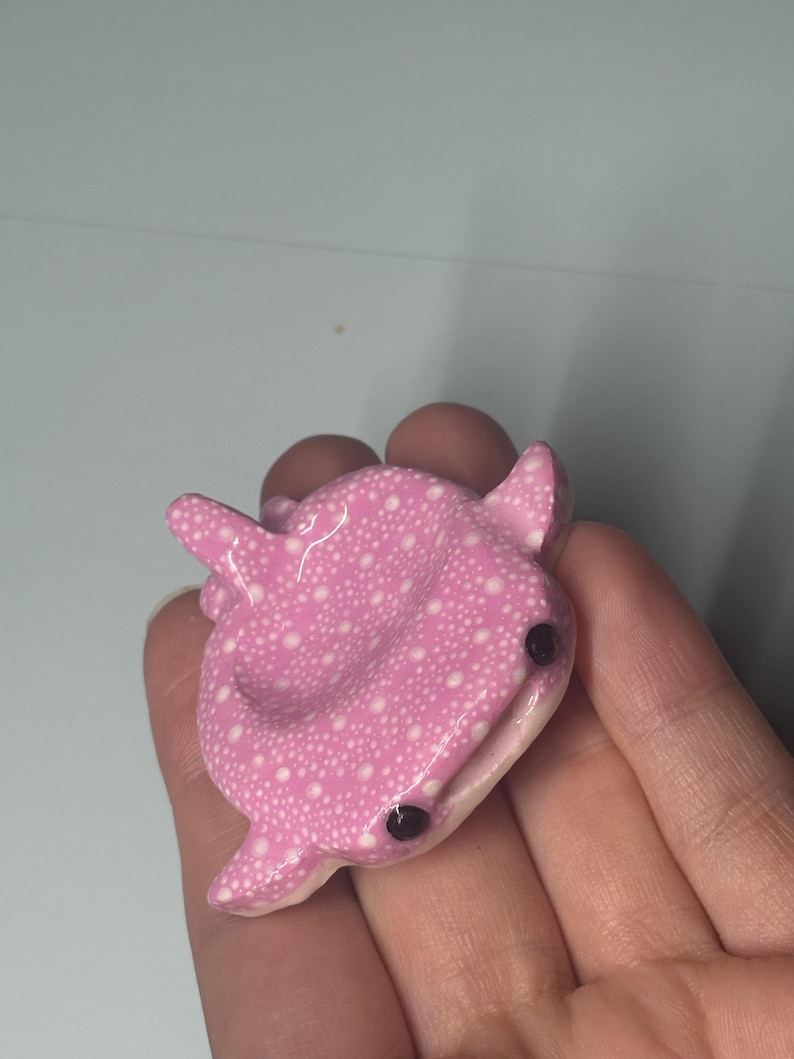 May include: A small, sculpted whale shark figurine featuring a glossy pink surface covered in a pattern of tiny white dots. The creature has two prominent black bead-like eyes and a wide, slightly open mouth, with fins extending from its sides.