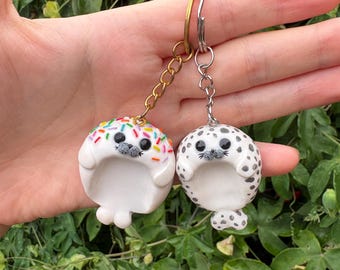 May include: Two round seal-shaped keychains made of white material. One seal features colorful sprinkles on its head and a gold-toned metal chain. The other seal has black spots across its body and a silver-toned metal chain. Both have black eyes.
