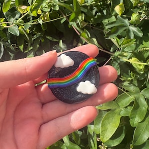 May include: A circular black pin with a glittery finish featuring a raised, colourful rainbow arching across the centre. Two small, white, puffy clouds are positioned on opposite sides of the rainbow, one at the top left and one at the bottom right.