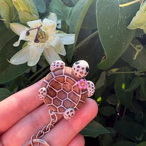 Può includere: Un portachiavi a forma di tartaruga con guscio marrone a motivi esagonali bianchi, un piccolo fiore rosa e quattro pinne maculate. La tartaruga ha la testa bianca con occhi scuri ed è fissata a una catenella in metallo argentato con anello.