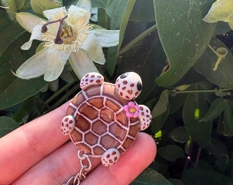 May include: A turtle keychain featuring a brown shell with white hexagonal patterns, a small pink flower accent, and four spotted flippers. The turtle has a white head with dark eyes and is attached to a silver metal chain and a circular key ring.