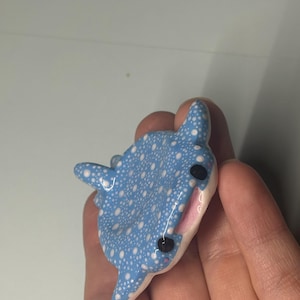 May include: A small, sculpted whale shark figurine featuring a light blue body covered in white polka dots. The figure has two black eyes, a white underbelly, and a small open mouth showing a pink interior, crafted with a smooth, glossy finish.