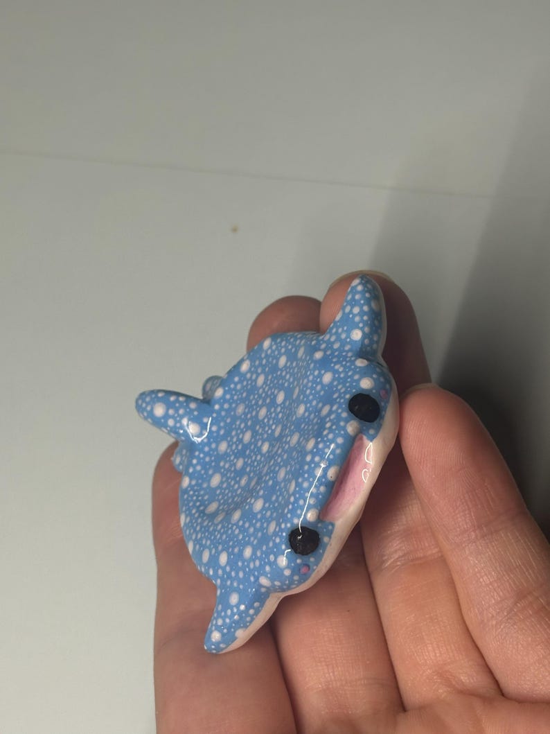 May include: A small, sculpted whale shark figurine featuring a light blue body covered in white polka dots. The figure has two black eyes, a white underbelly, and a small open mouth showing a pink interior, crafted with a smooth, glossy finish.