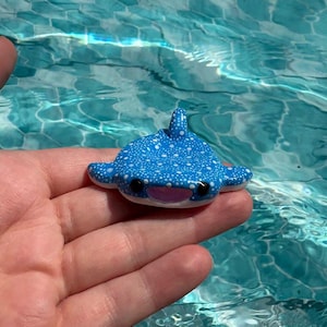 May include: A small, blue whale shark figurine featuring a pattern of white dots across its back, a dorsal fin, two side fins, a light pink mouth, and two black eyes. The figurine is shaped like a manta ray with a wide, flat body design.