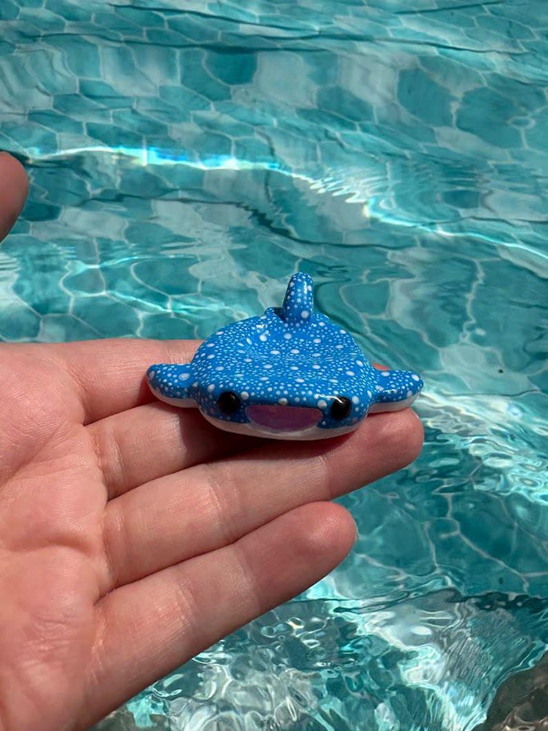 May include: A small, blue whale shark figurine featuring a pattern of white dots across its back, a dorsal fin, two side fins, a light pink mouth, and two black eyes. The figurine is shaped like a manta ray with a wide, flat body design.
