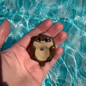 Könnte beinhalten: Ein braun-cremefarbener Otter-Anhänger, der einen kleinen grauen Stein auf seinem Bauch hält. Der Otter hat schwarze Augen, eine rosa Nase und einen silbernen Metallring an der Oberseite zum Aufhängen. Der Anhänger hat eine glatte und glänzende Oberfläche.