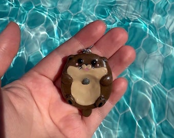 May include: A brown and cream-colored otter charm featuring a small gray stone held on its belly. The otter has black eyes, a pink nose, and a silver metal jump ring attached to the top for hanging. The charm is smooth and glossy in appearance.